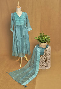 Teal embroidered dress on a mannequin with a plant on a woven stool against a brown background