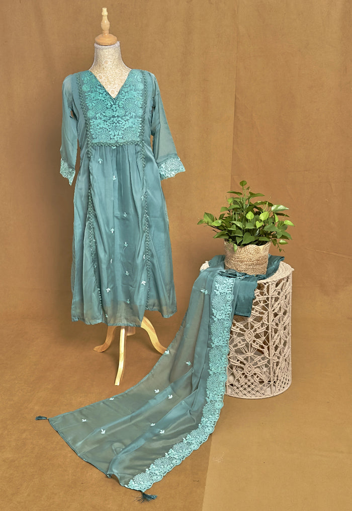 Teal embroidered dress on a mannequin with a plant on a woven stool against a brown background