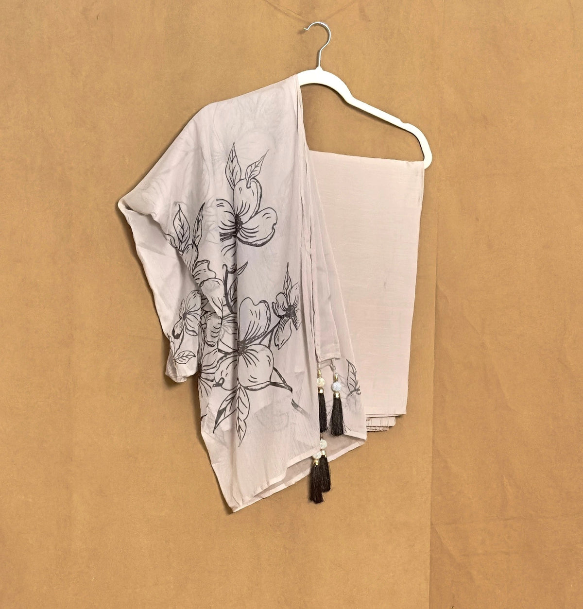 White scarf with black floral patterns and tassels on a hanger against a beige background