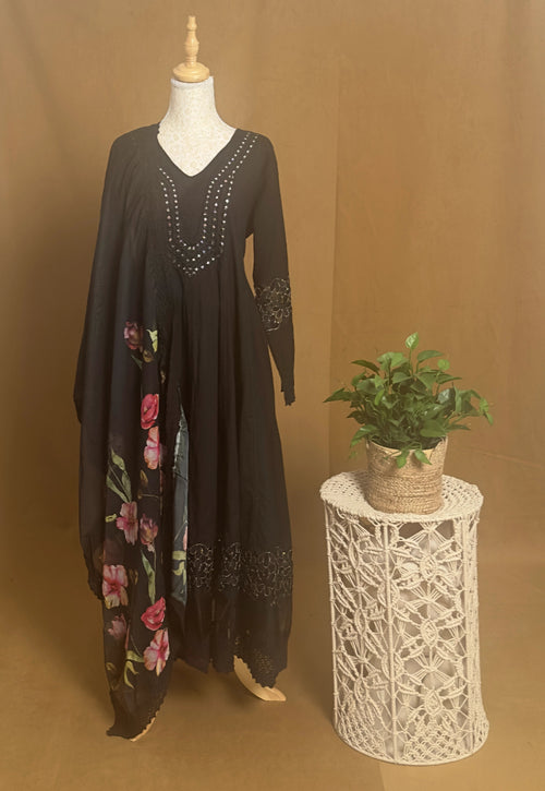 Black dress with floral embroidery on a mannequin next to a small plant on a lace stool against a brown background