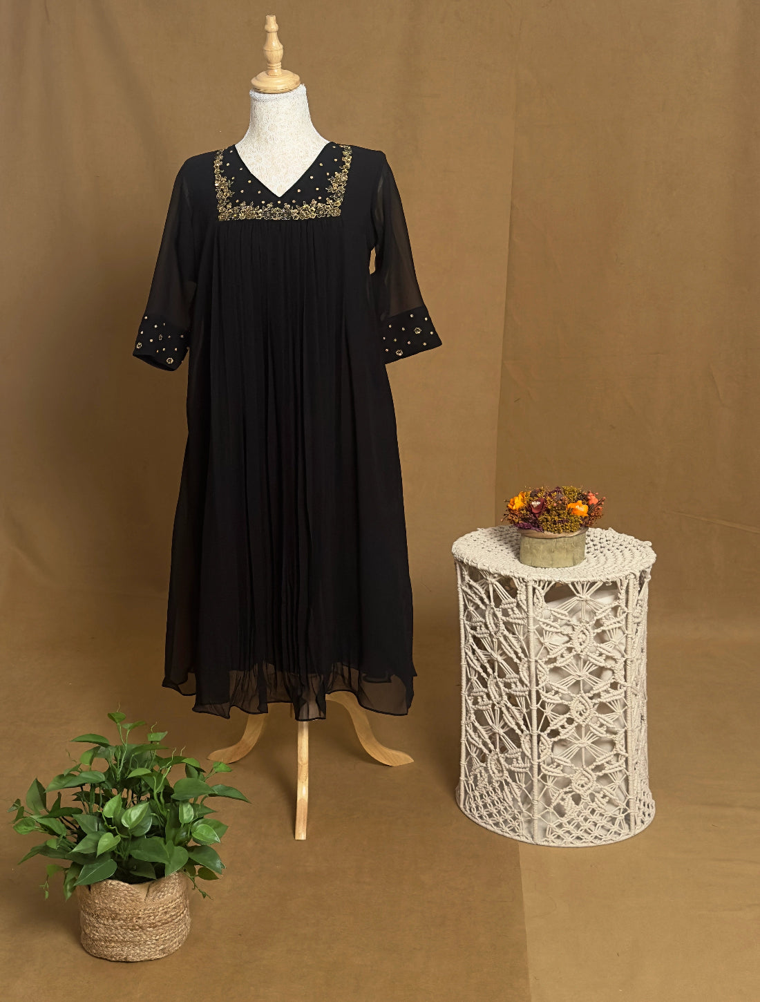 Black dress on a mannequin with decorative embroidery, placed next to a small table with a plant and decorative items against a brown background.
