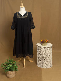 Black dress on a mannequin with decorative embroidery, placed next to a small table with a plant and decorative items against a brown background.