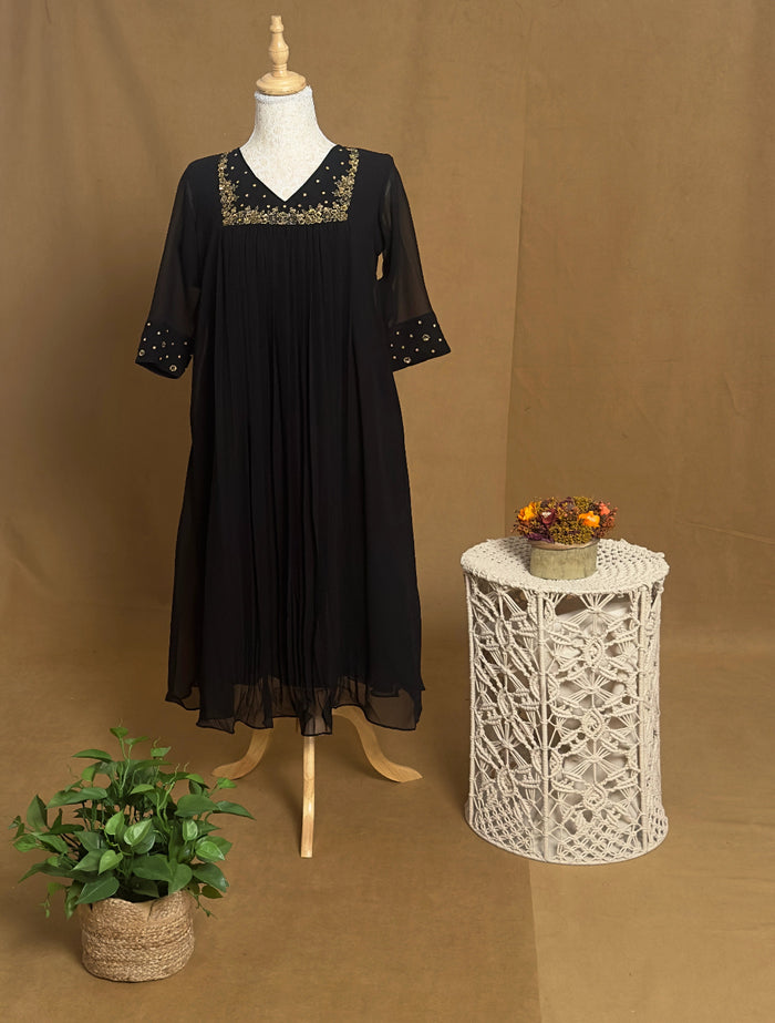 Black dress on a mannequin with decorative embroidery, placed next to a small table with a plant and decorative items against a brown background.
