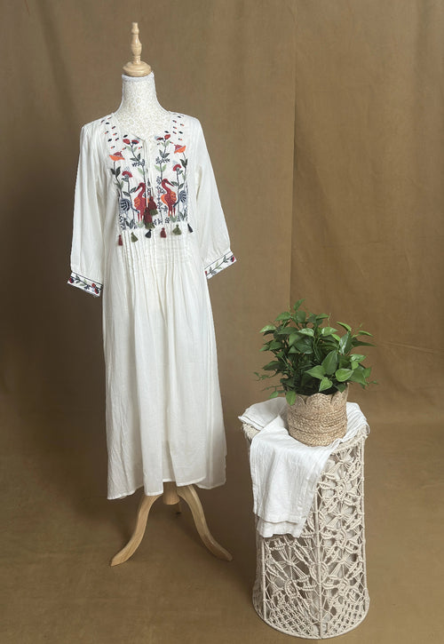 White embroidered dress on a mannequin with a small table and plant against a brown background