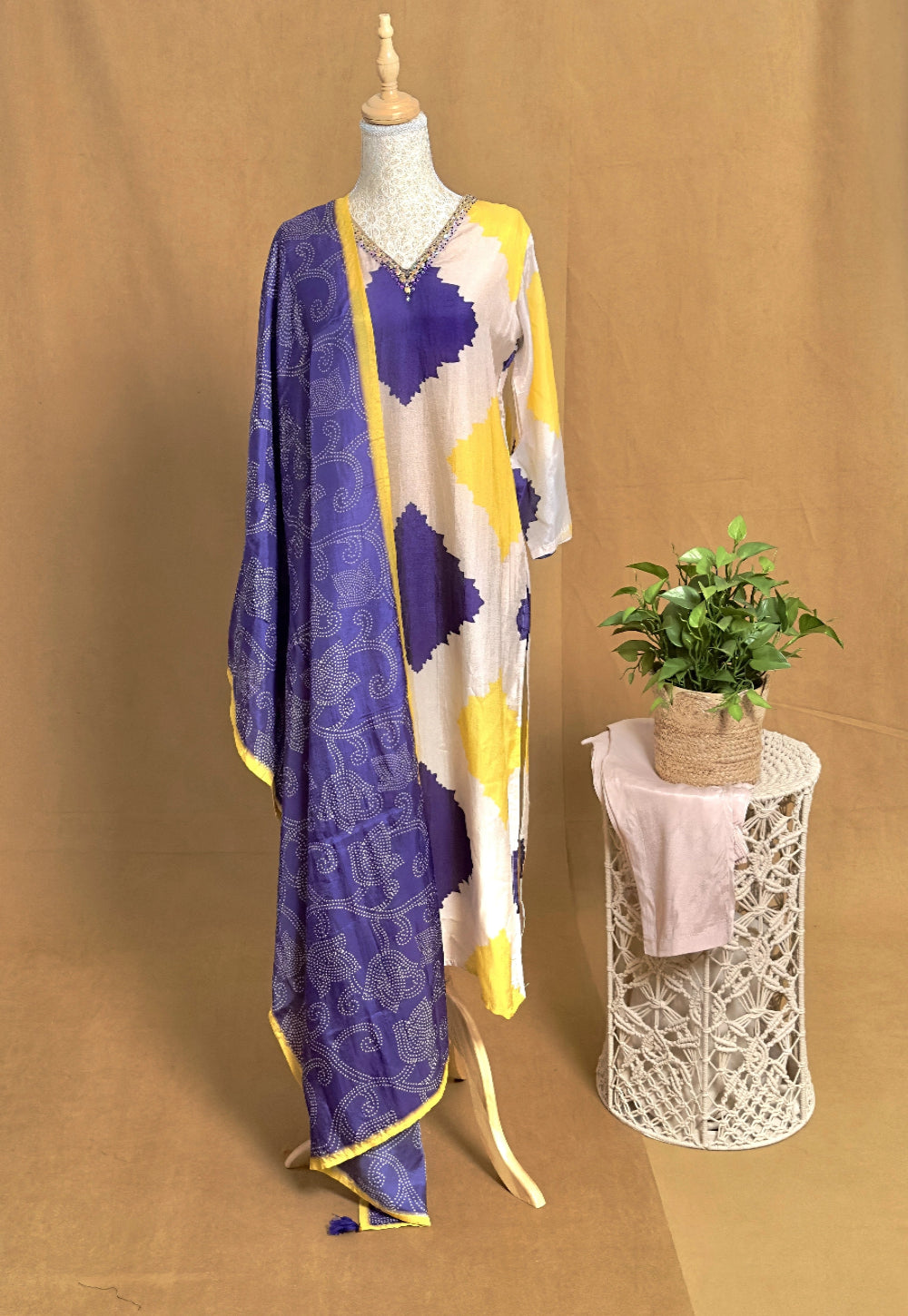 Mannequin wearing a purple and yellow patterned scarf with a plant on a stand against a brown background