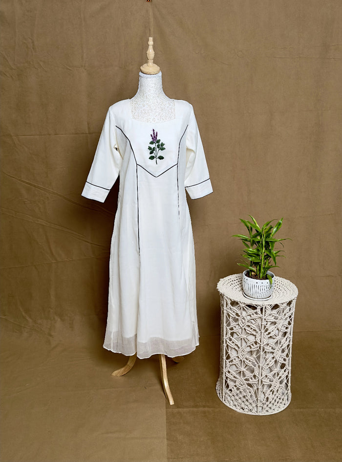 White dress with embroidery on a mannequin next to a small table with a plant against a brown background